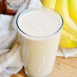 Pint glass with skinny almond banana breakfast smoothie on a wood table with a spoon and peanut butter at the bottom left, bananas and sweet n low in the background to the right and a white cloth and jar of peanut butter in the background to the left