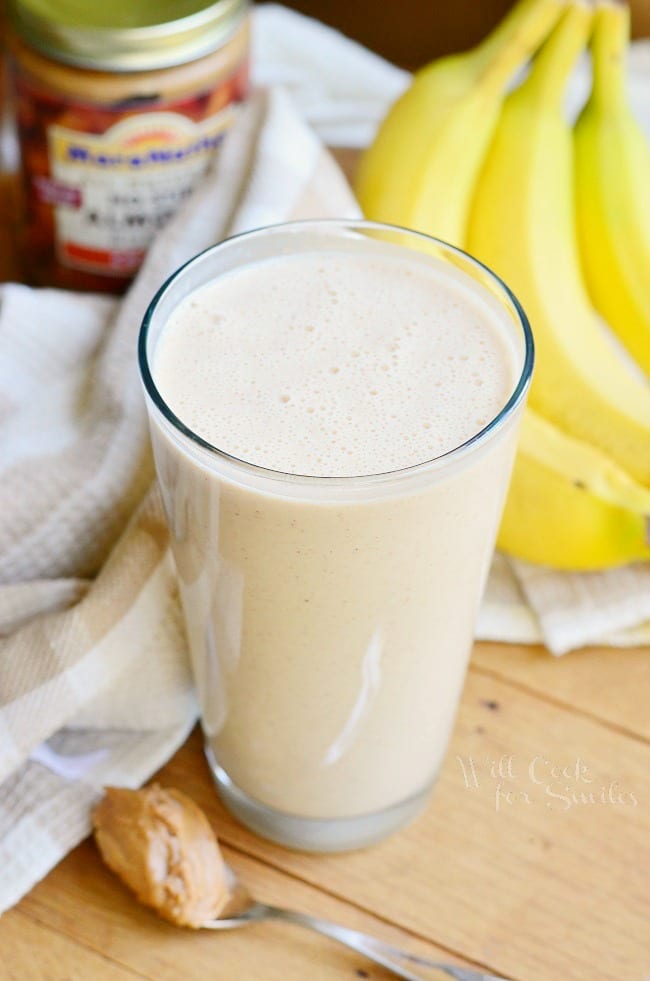 Pint glass with skinny almond banana breakfast smoothie on a wood table with a spoon and peanut butter at the bottom left, bananas and sweet n low in the background to the right and a white cloth and jar of peanut butter in the background to the left