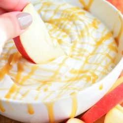 oval dish with skinny caramel cream dip with apples scattered around the dish and 1 whole apple in the background to the right and a hand dippingan apple slice into the dip