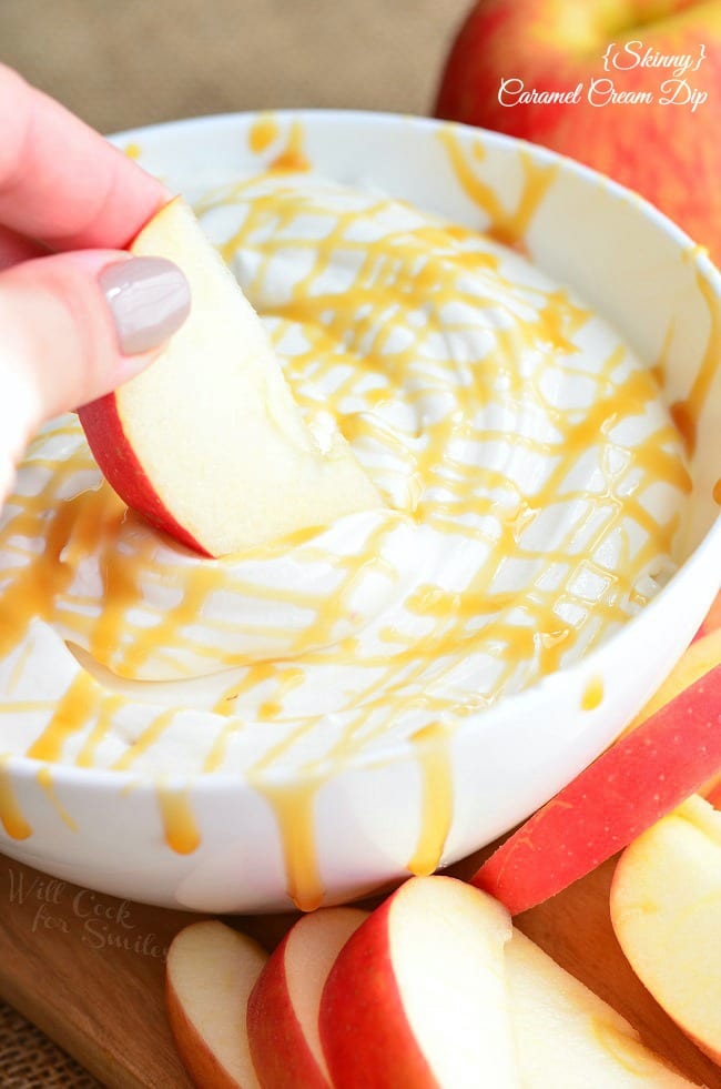 oval dish with skinny caramel cream dip with apples scattered around the dish and 1 whole apple in the background to the right and a hand dippingan apple slice into the dip