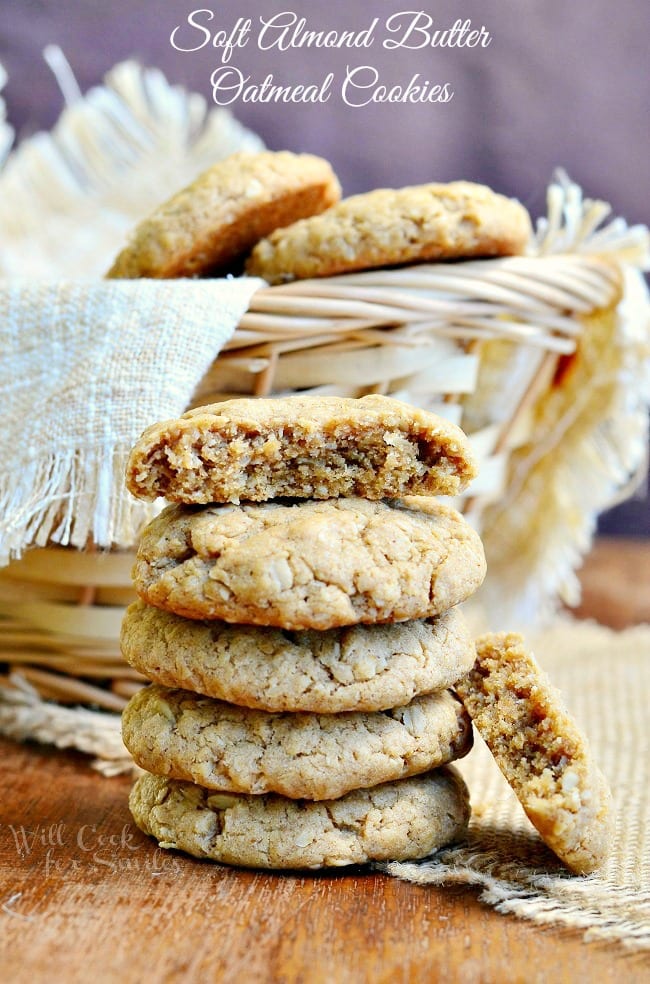 Soft Almond Butter Oatmeal Cookies stacked up on a table with a basket in the background with cookies in it  with the top one cut in half 