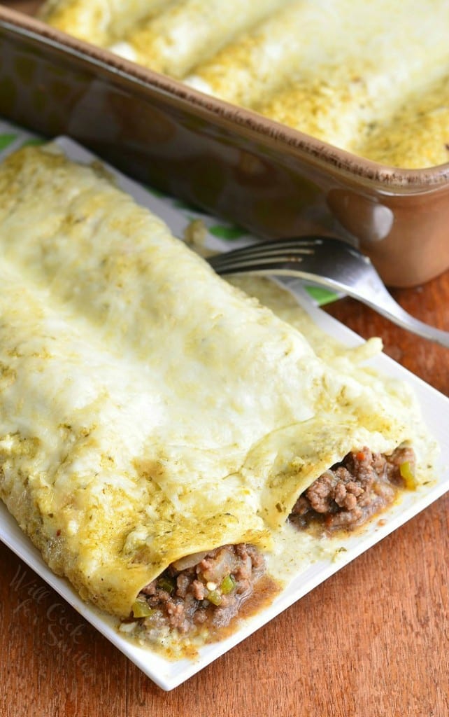small rectangular plate with 2 portions of beef enchiladas on a wooden table with a fork resting between the plate and a baking pan in the background with additional portions of beef enchiladas as viewed from above