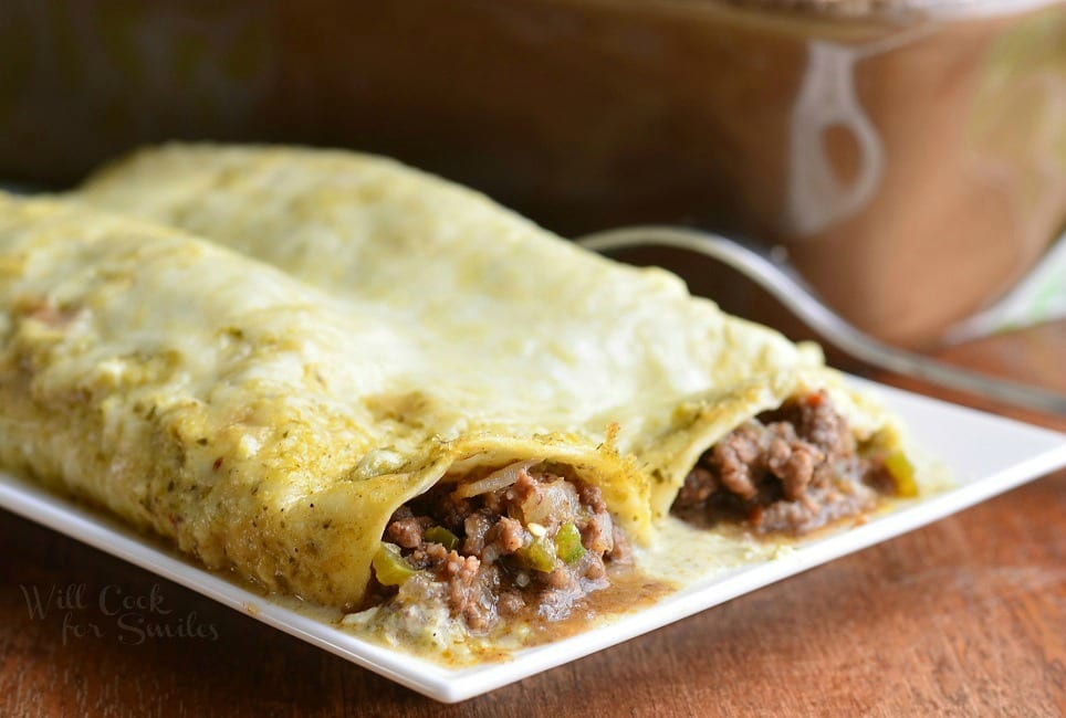 close up view of small rectangular plate with 2 portions of beef enchiladas on a wooden table with a fork resting between the plate and a baking pan in the background with additional portions of beef enchiladas.