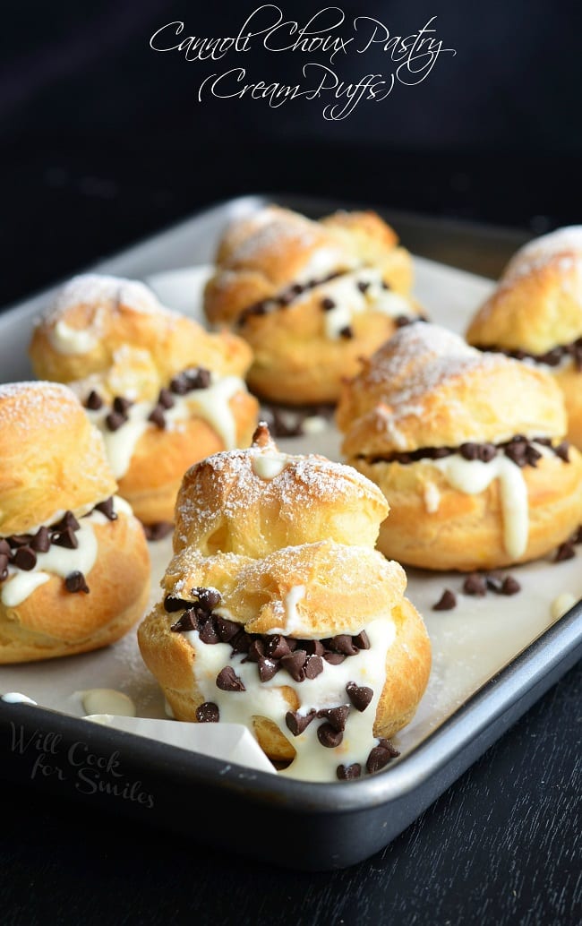 Cannoli Choux Pastry (Cream Puffs) are lined in a pan. Delicate pâte à choux pastries are filled with Cannoli cream. It's also topped with some mini chocolate morsels and powder sugar.