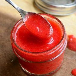 small mason jar filled with easy strawberry sauce on a wooden table with a spoon pulling some sauce from jar and the mason lid jar in the background