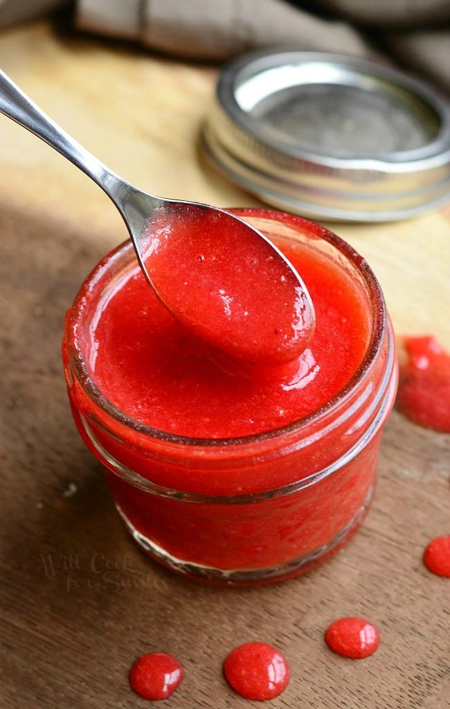 small mason jar filled with easy strawberry sauce on a wooden table with a spoon pulling some sauce from jar and the mason lid jar in the background