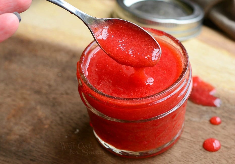 Easy Strawberry Sauce in a small mason jar with a spoon scooping some out on a cutting board with little drops of sauce around it 