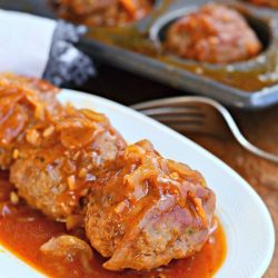 small white oval dish with french onion baked meatballs on a wooden table with a fork and cupcake baking pan filled with additional meatballs in it in background.