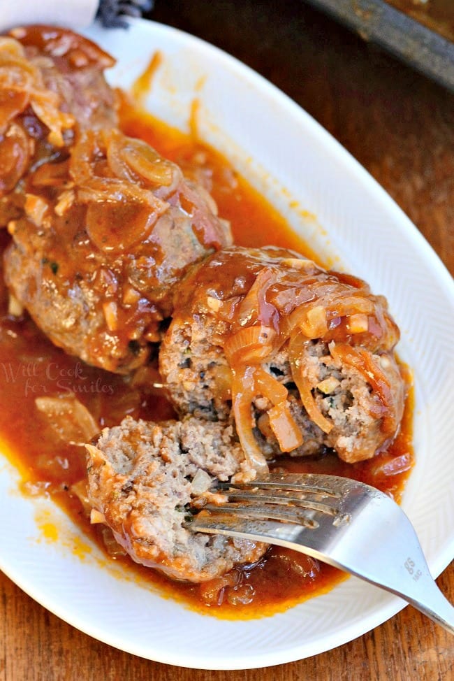 small white oval dish with french onion baked meatballs on a wooden table with a fork splitting the first meatball and taking one bite