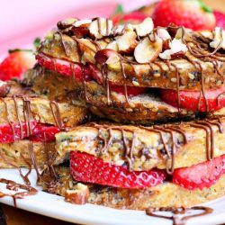 Small round white plate with Fruit filled hazelnut french toast with chocolate drizzle topping on a wooden table with a fork in the foreground and a pink cloth in the background to the left