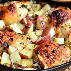 view from above of black skillet with one pot balsamic chicken thighs and potatoes on a tan burlap placemat with a wooden spoon resting on skillet.