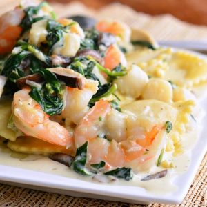 White decorative plate with ravioli with seafood spinach mushrooms in garlic sauce on a tan placemat on a wooden table with a spoon in the background.