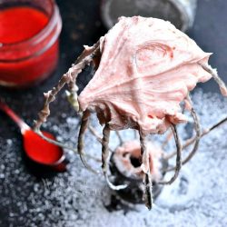 mixer attachment standing on a black table covered in powdered sugar while the attachment is covered in strawberry buttercream frosting as viewed from above