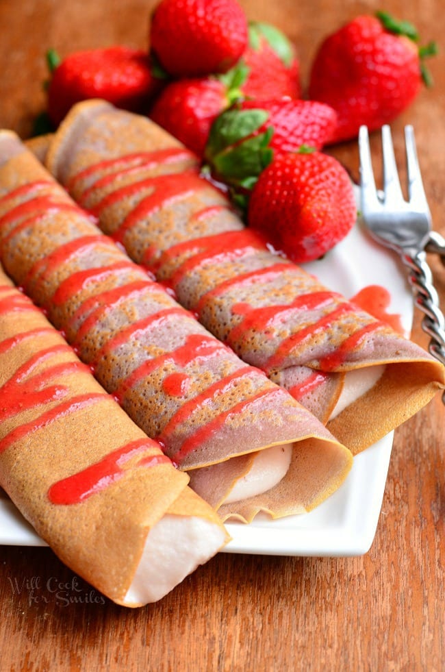 white decorative plate with strawberry crepes and strawberry mascarpone with drizzled strawberry sauce and strawberries on the plate in the background. All sit on a wooden table as viewed from above
