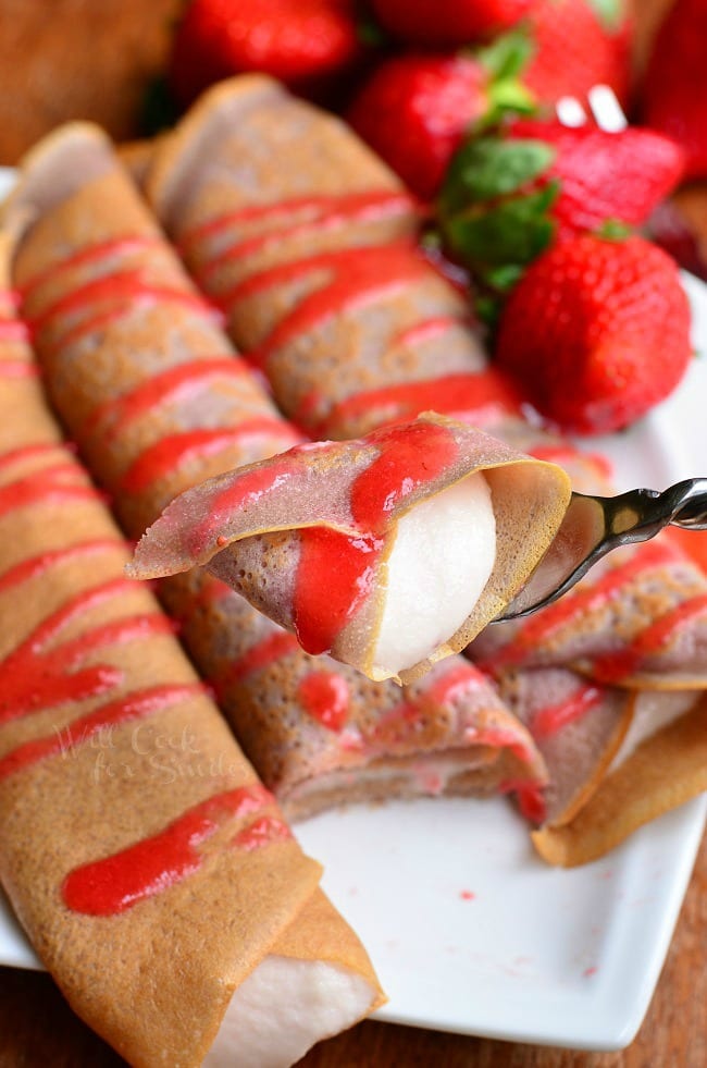 white decorative plate with strawberry crepes and strawberry mascarpone with drizzled strawberry sauce and strawberries on the plate in the background with a fork holding one bite above the plate