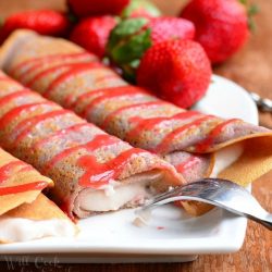 white decorative plate with strawberry crepes and strawberry mascarpone with drizzled strawberry sauce and strawberries on the plate in the background. All sit on a wooden table.