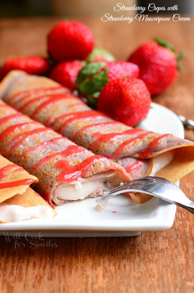 white decorative plate with strawberry crepes and strawberry mascarpone with drizzled strawberry sauce and strawberries on the plate in the background. All sit on a wooden table.