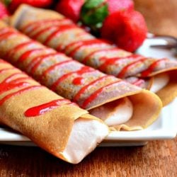 a close up view of white decorative plate with strawberry crepes and strawberry mascarpone with drizzled strawberry sauce and strawberries on the plate in the background. All sit on a wooden table.