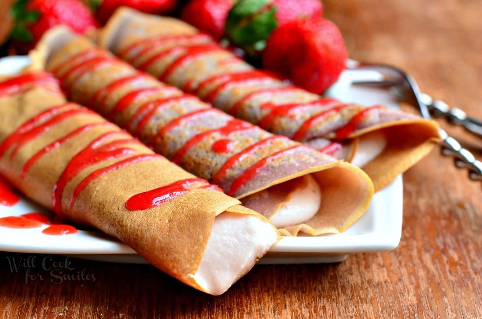 a close up view of white decorative plate with strawberry crepes and strawberry mascarpone with drizzled strawberry sauce and strawberries on the plate in the background. All sit on a wooden table.