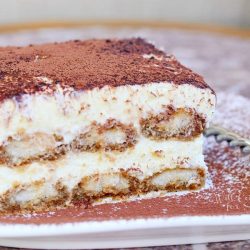 white square decorative plate dusted with cocoa and a slice of tiramisu. A fork resting on the plate to the right side of the slice all on a placemat and a wooden table as viewed close up