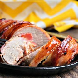 close up view of Bacon wrapped garlic dijun pork loin sliced on a black skillet on a wooden table with a yellow and white cloth in the background