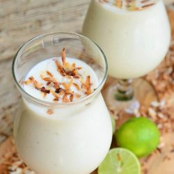 2 dessert beverage glasses filled with creamy tropical smoothie on a wooden cutting board for presentation and toasted coconut flakes and sliced limes scattered around the base of the glasses.