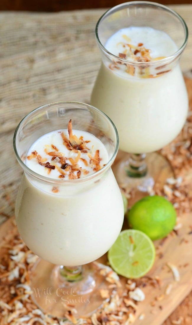 2 dessert beverage glasses filled with creamy tropical smoothie on a wooden cutting board for presentation and toasted coconut flakes and sliced limes scattered around the base of the glasses.
