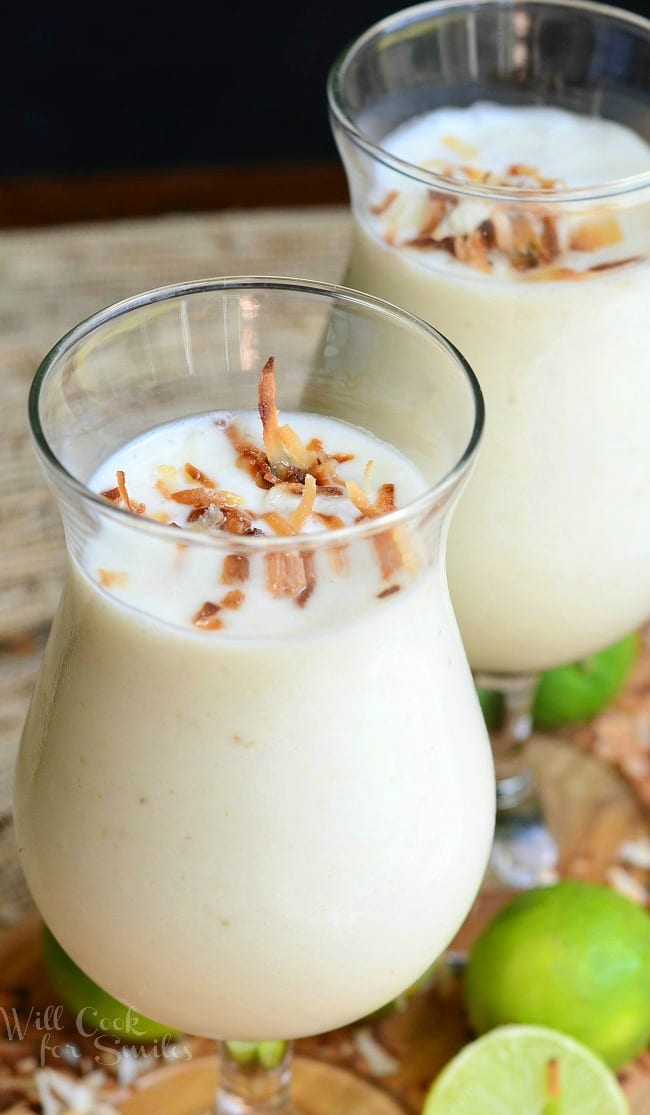 Smoothies in two glasses with toasted coconut on top on a cutting board with toasted coconut and limes around it 