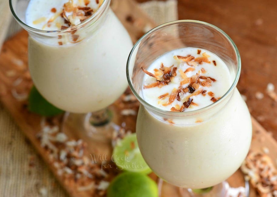 top view of Smoothies in two glasses with toasted coconut on top on a cutting board with toasted coconut and limes around it 