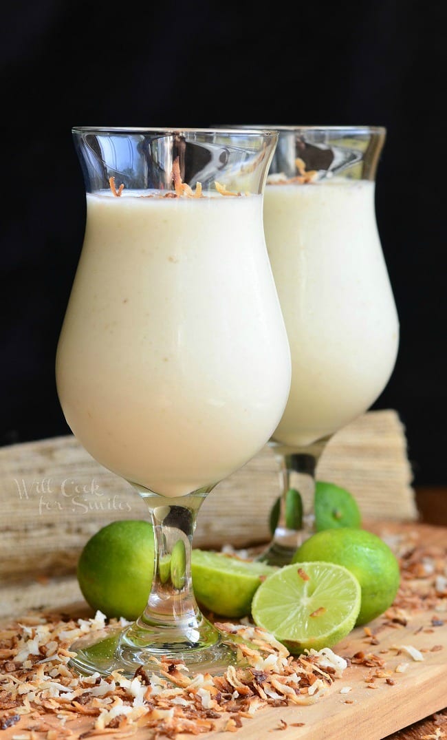 Smoothies in two glasses with toasted coconut on top on a cutting board with toasted coconut and limes around it 