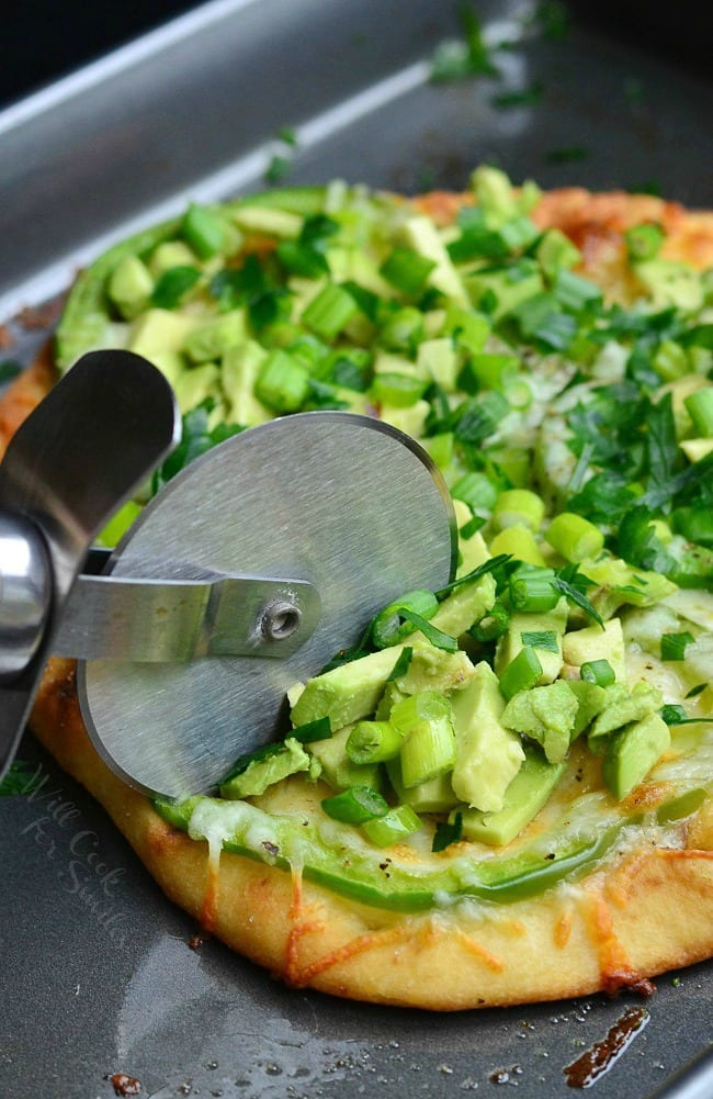 slicing Green Naan Pizza with avocado, green peppers and green onions on top with a pizza slicer 