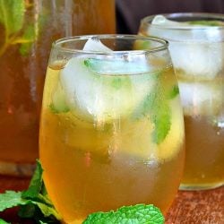 2 glasses of brewed honey mint green iced tea on a wooden table with mint laying on the table to the left of the glasses and a pitcher filled with more tea in the background to the left of glasses.