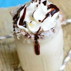 view from above of mason jar filled with lightened almond joy milkshake on a tan placemat with 3 straw around the base of the jar and a blue cloth in the background topped with whipped cream and chocolate syrup