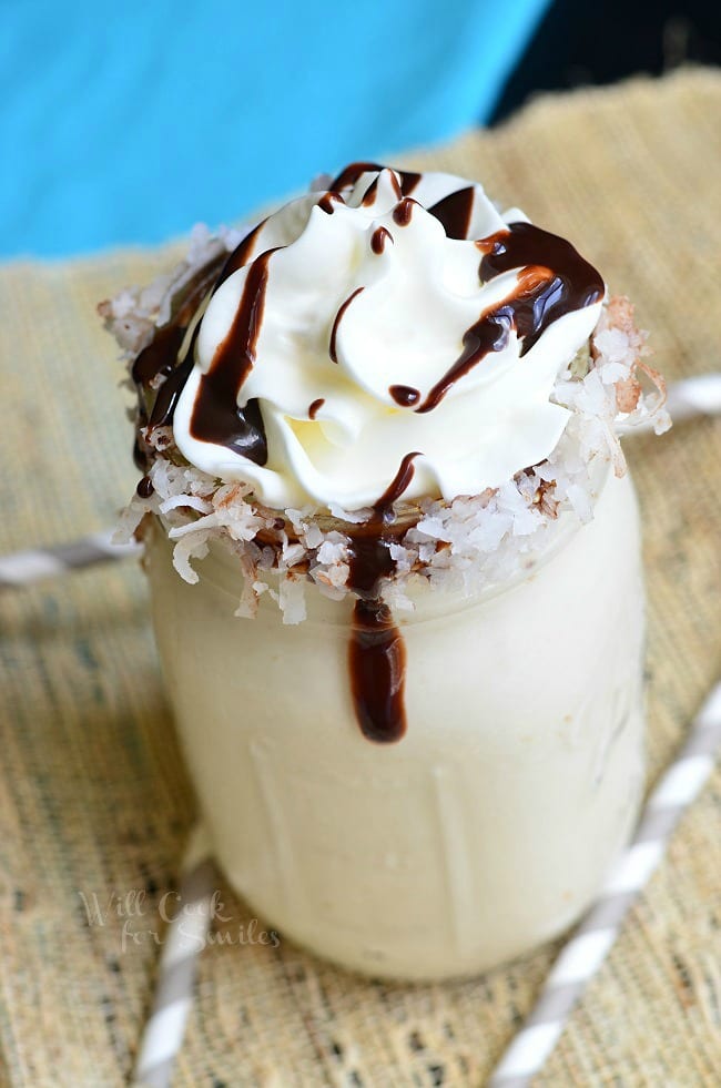 Milkshake in a mason jar with coconut around the rim and whip cream on top with a chocolate sauce drizzle over it 