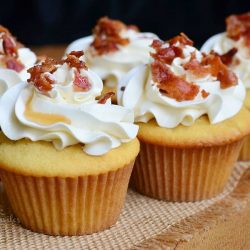 close up view of 4 maple bacon cupcakes on a burlap cloth on wooden table with a black background