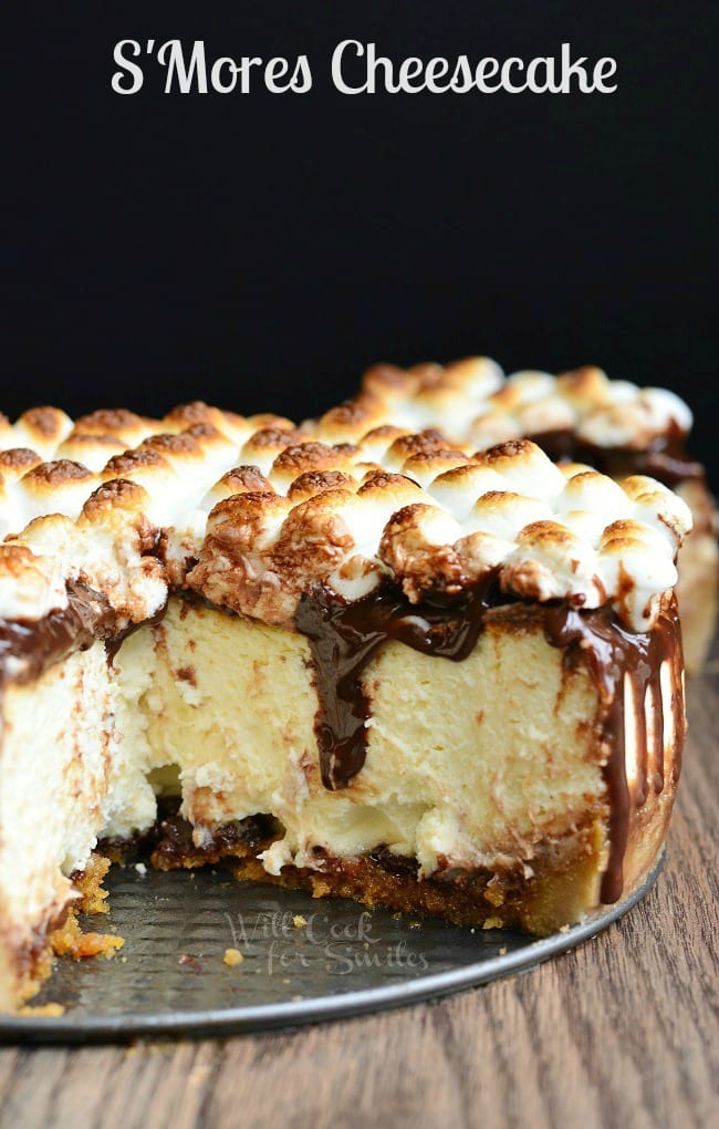 S'Mores Cheesecake with mini marshmallows on top with chocolate sauce over it with a piece of cheesecake missing 
