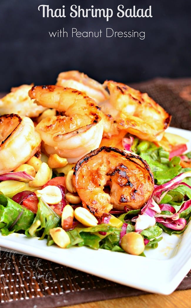 Thai Shrimp Salad with Peanut Dressing is served on a white plate. Plenty of plump, cooked shrimp are perched on a bed of greens. Another key component is the peanuts.