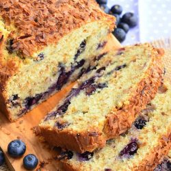 loaf of toasted coconut blueberry bread with the first 3 slices stacked in front of the rest of the loaf on a wooden cutting board with blueberries scattered around on table.