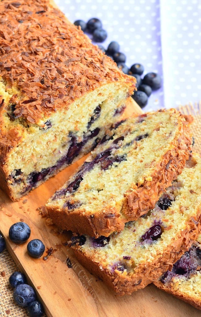 Toasted Coconut Blueberry Bread served on a wooden board. Coconut flakes are noticeable on the top of this loaf. The sliced pieces of bread show the blueberries within. There are also fresh blueberries laying on the board and table. 