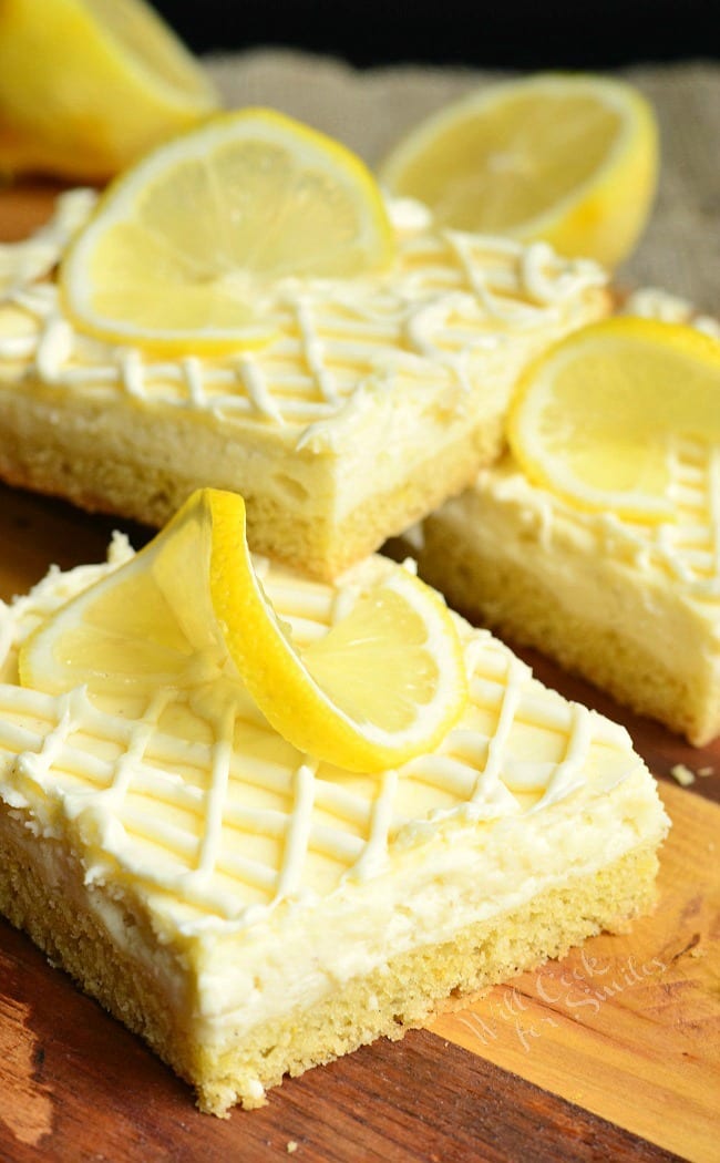 Vanilla Bean Lemon Cheesecake Bars lay piled on a wooden board. The piping work is detailed on top of each bar. Each one is also topped with a fresh lemon twist. Two lemon halves fade into the background.