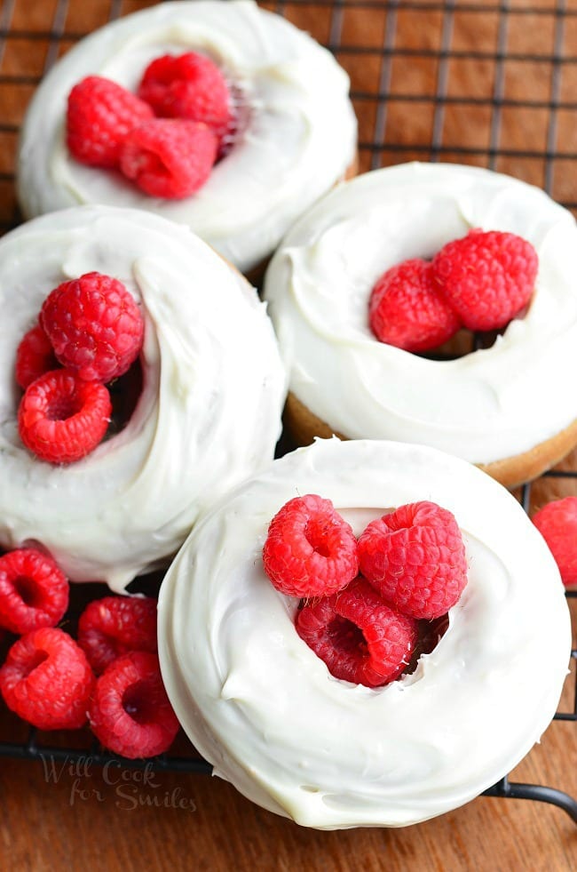 view from above of 4 white chocolate covered baked raspberry donuts on a wire cooling rack and whole raspberries laying at bottom left of donuts.