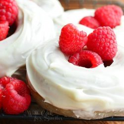 close up view of 4 white chocolate covered baked raspberry donuts on a wire cooling rack and whole raspberries laying at bottom left of donuts.