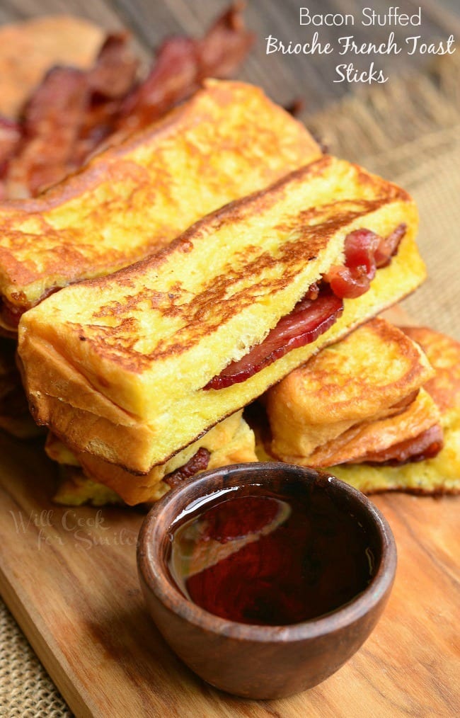 Bacon Stuffed Brioche French Toast Sticks stacked on a wooden board. In front of the sticks is a tiny bowl of maple syrup for dipping.