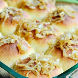 close up view of glass baking pan filled with caramelized onion dinner rolls topped with onions on a wooden table with a green cloth behind