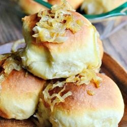 close up view of small wooden plate with 3 caramelized onion dinner rolls with a glass baking dish and more rolls in the background all on a wooden table