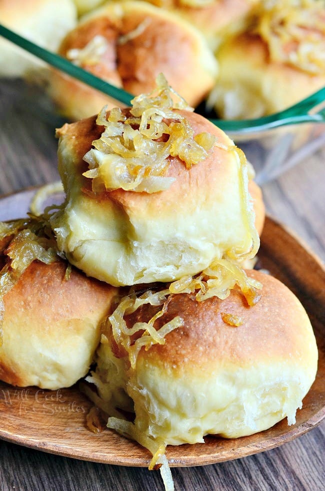 close up view of small wooden plate with 3 caramelized onion dinner rolls with a glass baking dish and more rolls in the background all on a wooden table