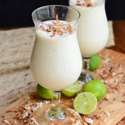 2 dessert glasses filled with creamy tropical smoothie on a wooden cutting board with coconut chavings around base of glasses along with cut limes