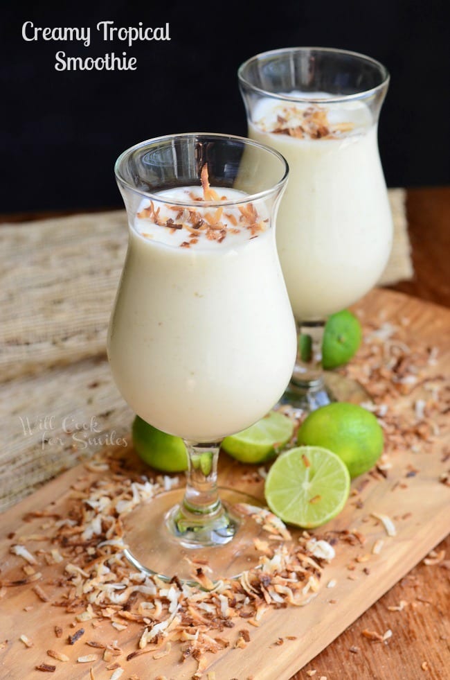 2 dessert glasses filled with creamy tropical smoothie on a wooden cutting board with coconut chavings around base of glasses along with cut limes