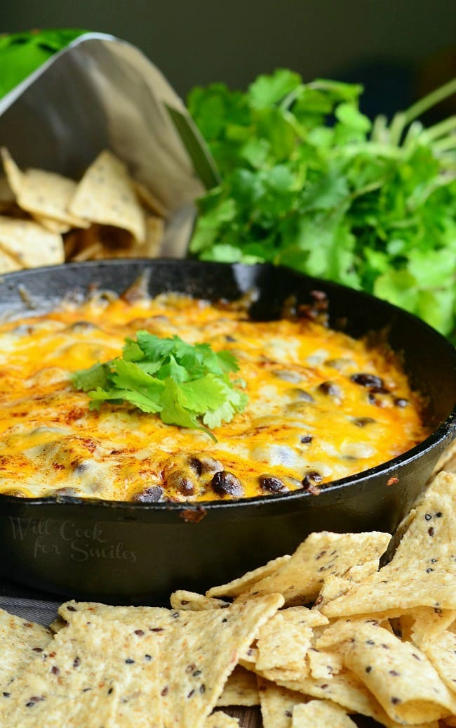 Hot 7 Layer Dip is presented here in a black skillet. It is topped with melted cheese and fresh cilantro. In the background is a bag of open tortilla chips and more cilantro. Tortilla chips are also placed around the front of the skillet.