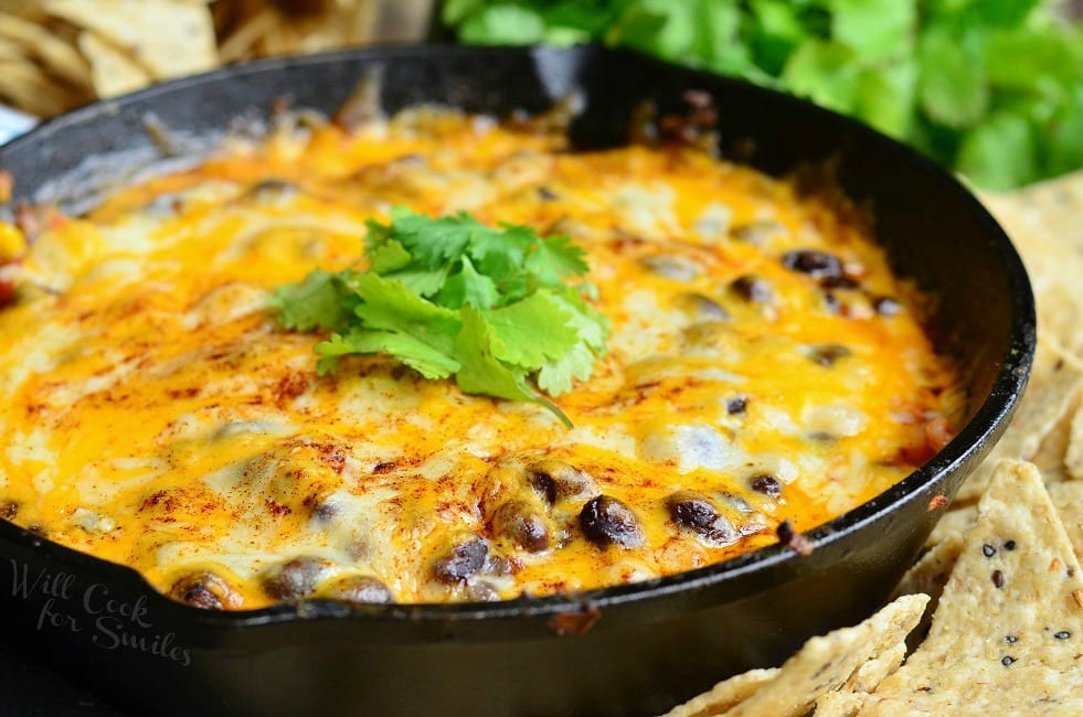 Hot 7 Layer Dip with cheese and black beans in a cast iron skillet with cilantro on top as garnish and tortilla chips around the pan 